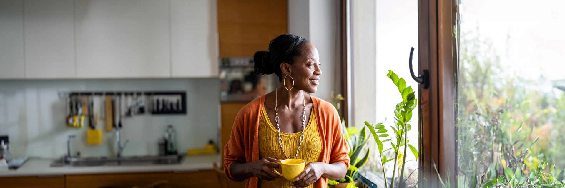 Middle-aged woman smiling and looking out of her kitchen window holding a cup of coffee